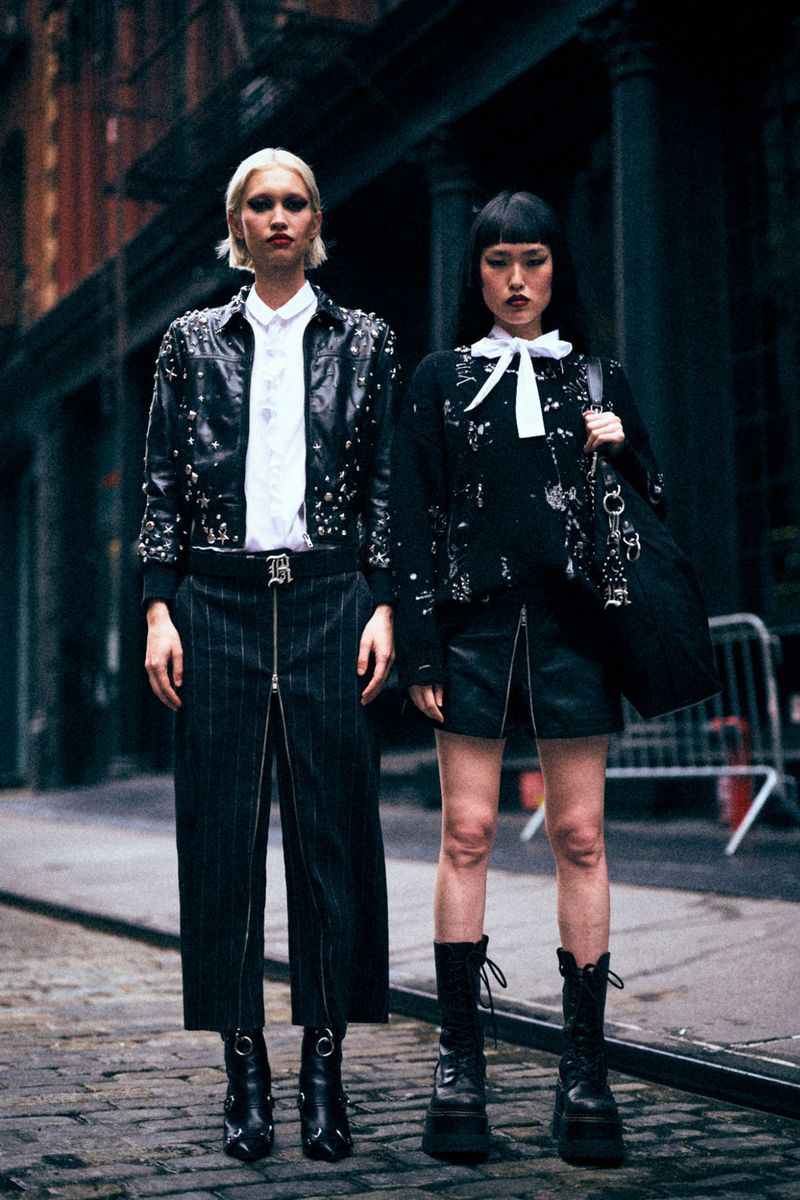 Two women in black, studded outfits stand on a city street. One has short blonde hair and pants with a full zipper. The other has black boots.