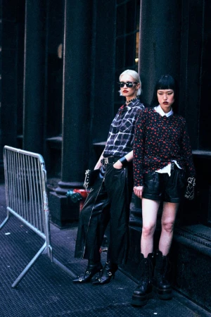 Two young women in dark, edgy fashion pose on an urban street with metal barriers; both have pale skin, dark clothing, and striking style.