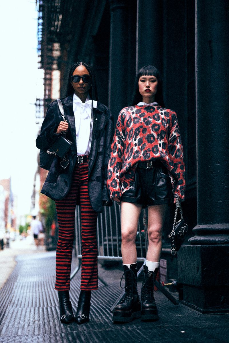 Two women in fashionable clothing pose in an urban setting with red striped pants, a leopard print sweater, and platform boots.