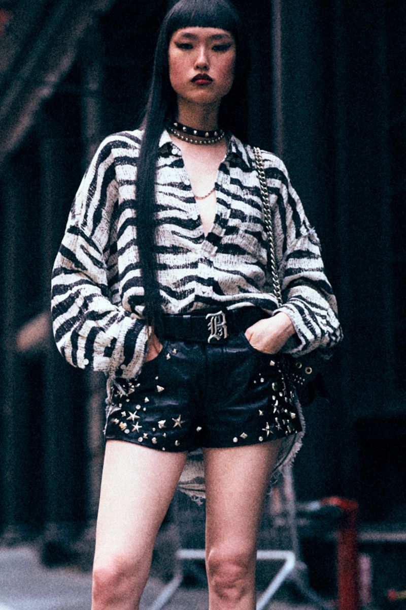 Asian woman in black studded shorts and animal-print shirt, posing in an urban setting with a modern, edgy fashion style.