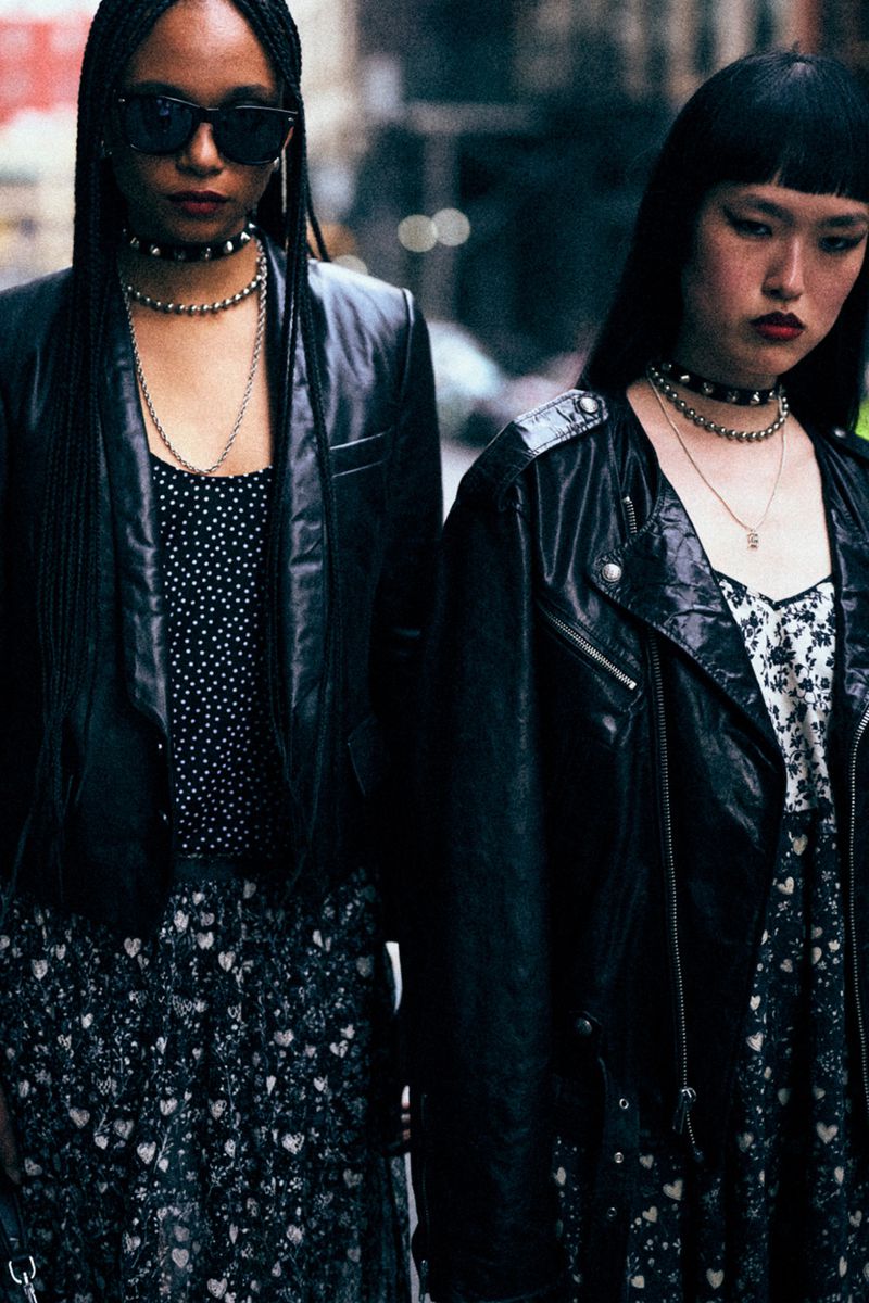 Two women stand outdoors in black leather jackets, floral print dresses, and sunglasses. They are wearing necklaces and chokers.