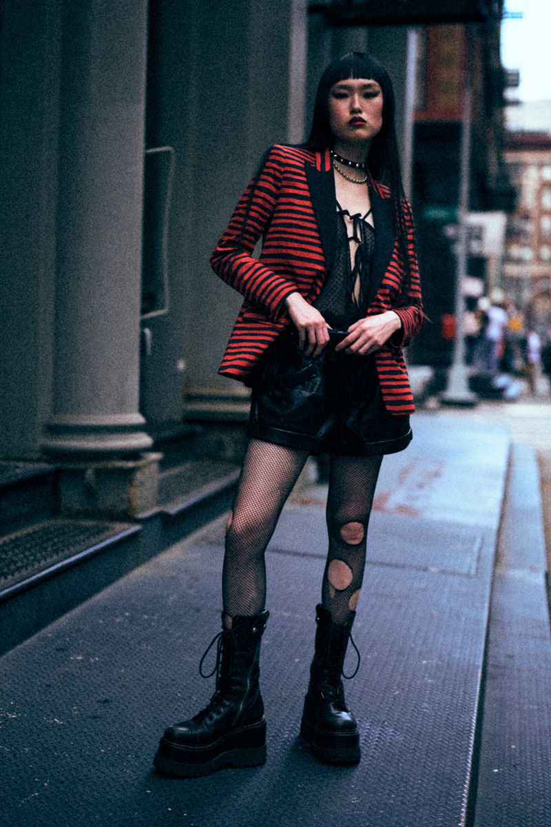 A woman in a red-striped blazer, black top, and shorts poses on a city street near grey stone architecture, showcasing an alternative fashion style.