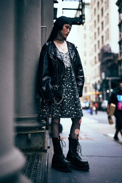 A woman in a black leather jacket, patterned dress, and platform boots stands on a city street next to a gray stone building pillar.