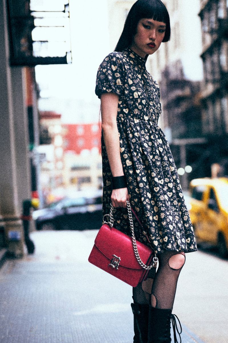 Asian woman in a floral dress standing in urban setting with a red purse and black boots, city street with buildings in the background.