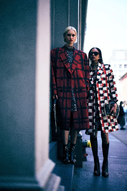Two women in patterned coats stand on a city street, one in a red and black plaid, the other in a black, white, and red pattern.