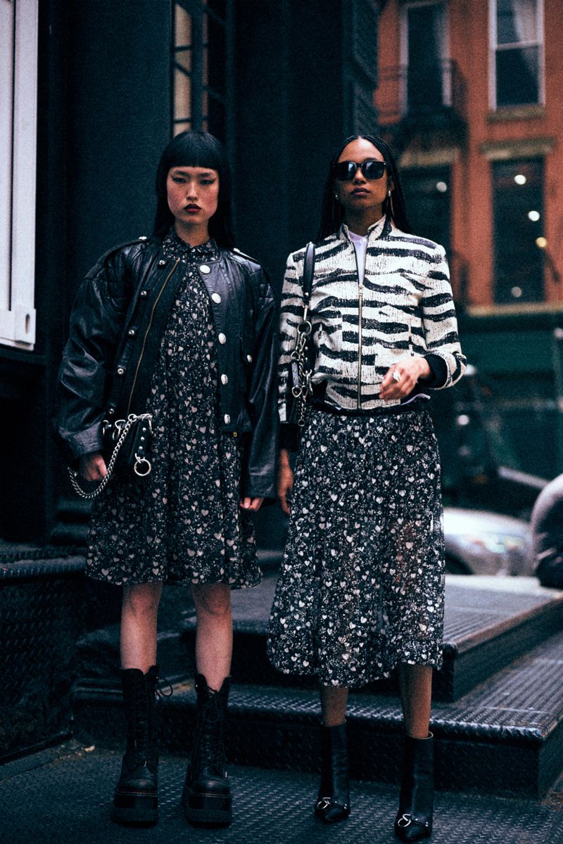 Two women in floral dresses and jackets pose on steps outdoors. One wears a leather jacket, the other a zebra print bomber.