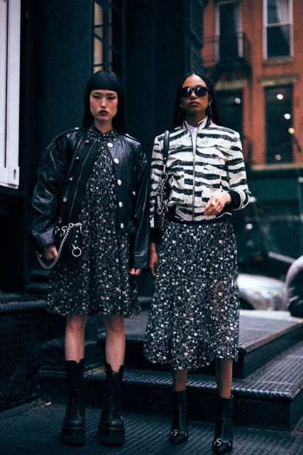 Two women in floral dresses and jackets pose on steps outdoors. One wears a leather jacket, the other a zebra print bomber.