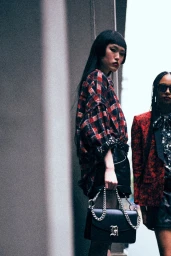 Model in red plaid shirt and black shorts holding a black chain-strap handbag standing beside another model outdoors