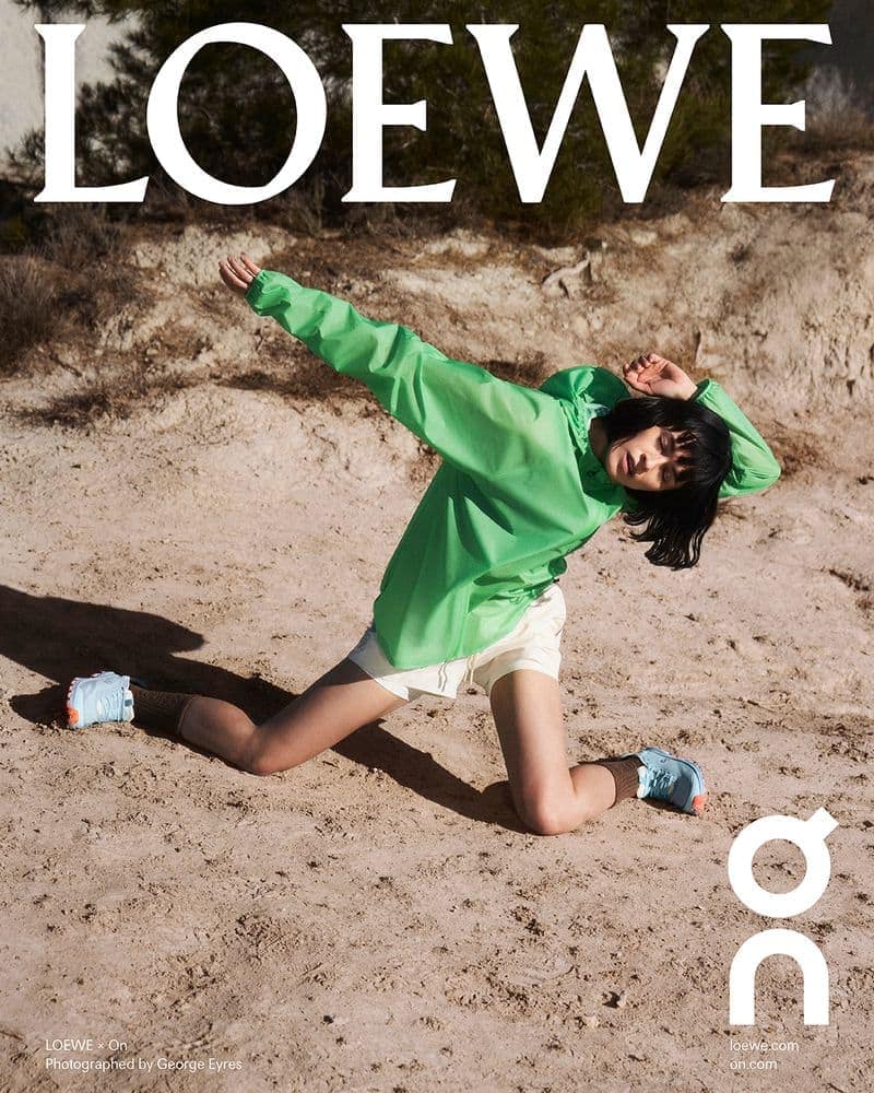 Model in green windbreaker and white shorts kneeling on sandy terrain under natural light for Loewe spring ad campaign