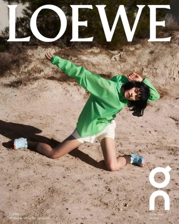 Model in green windbreaker and white shorts kneeling on sandy terrain under natural light for Loewe spring ad campaign