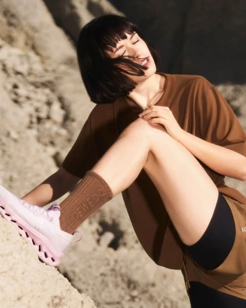 Woman in brown t-shirt and pink sneakers sitting outdoors against a sandy terrain background