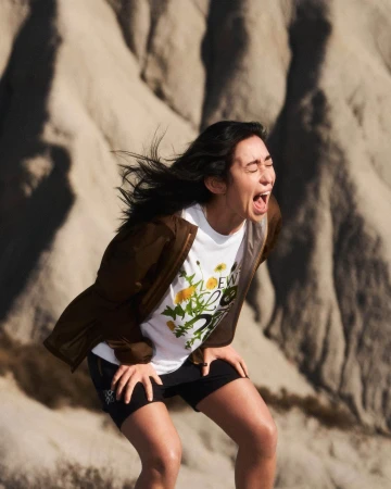 Woman in white t-shirt and brown jacket yelling outdoors against a barren sandy hillside background