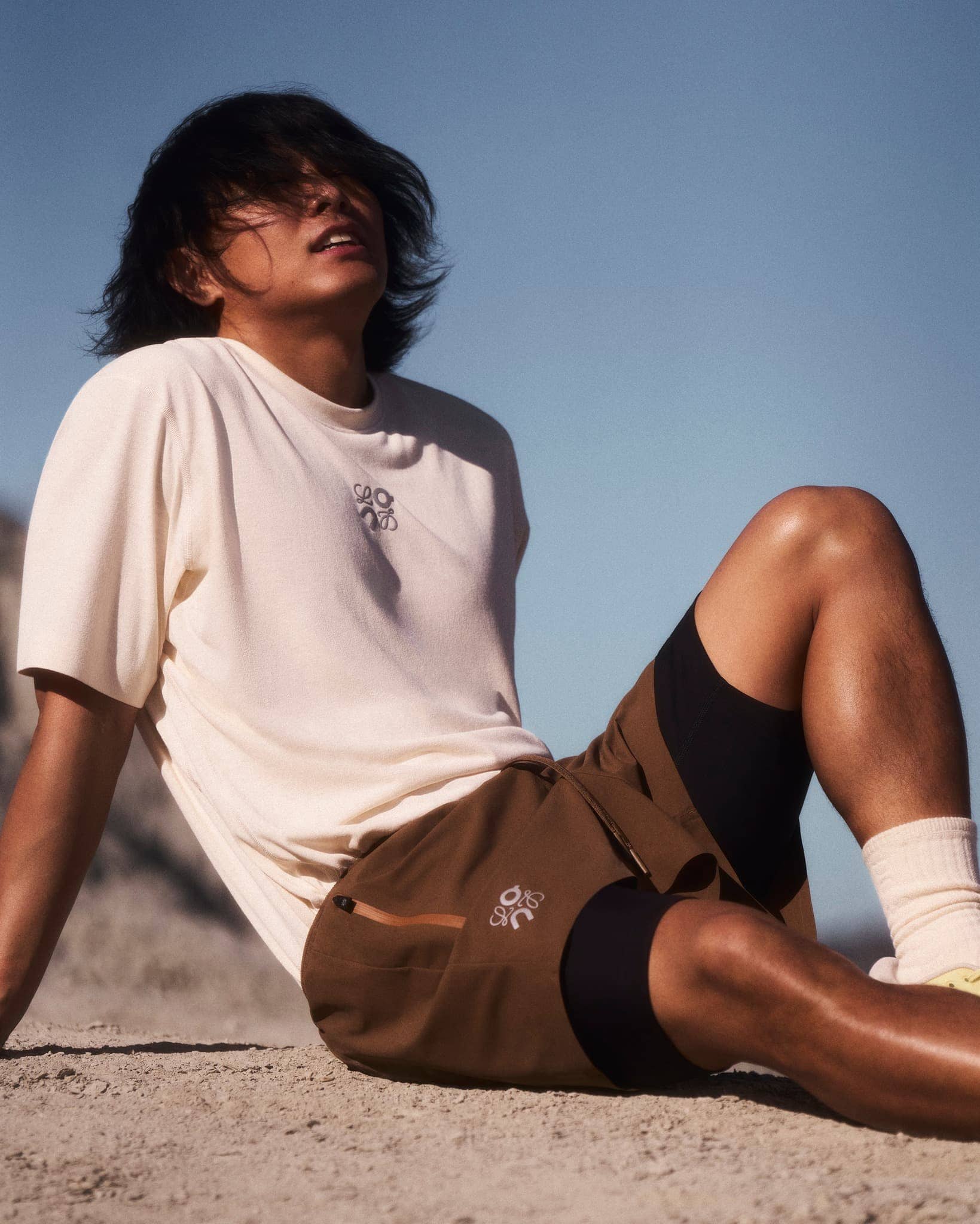 Man with dark hair seated on sandy ground wearing a cream t-shirt and brown shorts against a clear blue sky