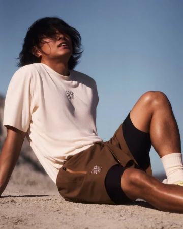 Man with dark hair seated on sandy ground wearing a cream t-shirt and brown shorts against a clear blue sky
