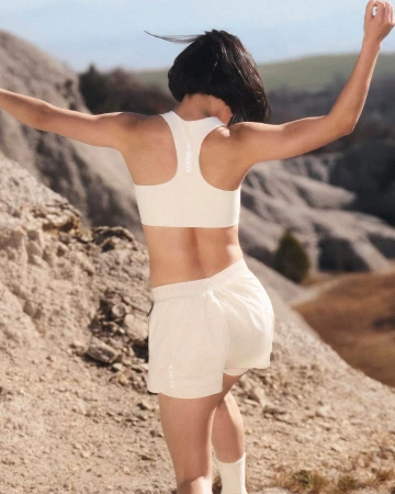 Woman in cream athletic wear running outdoors on a rocky hillside path with arms raised