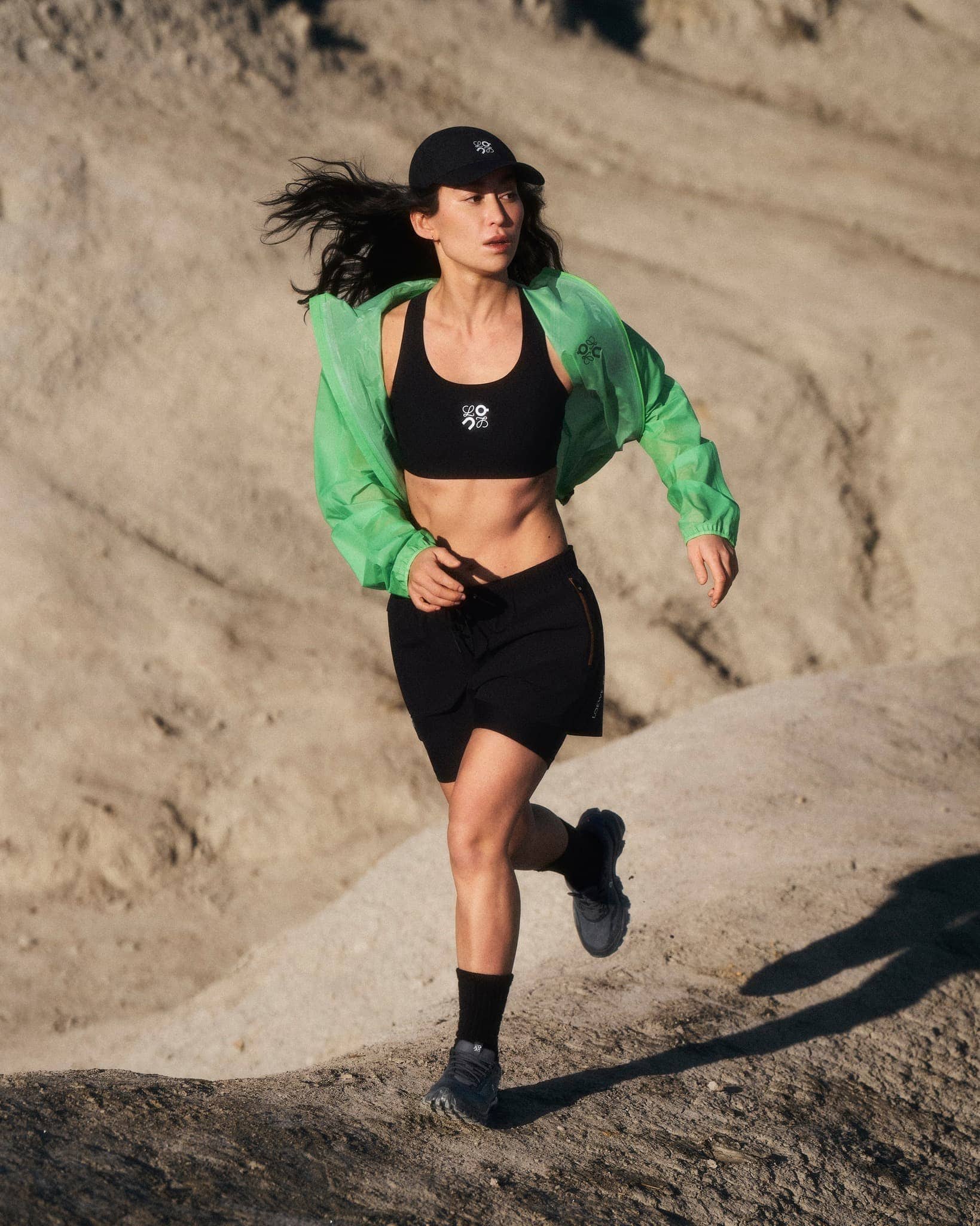Woman running outdoors on rocky terrain wearing a black sports bra, black shorts, and a vibrant green windbreaker jacket.