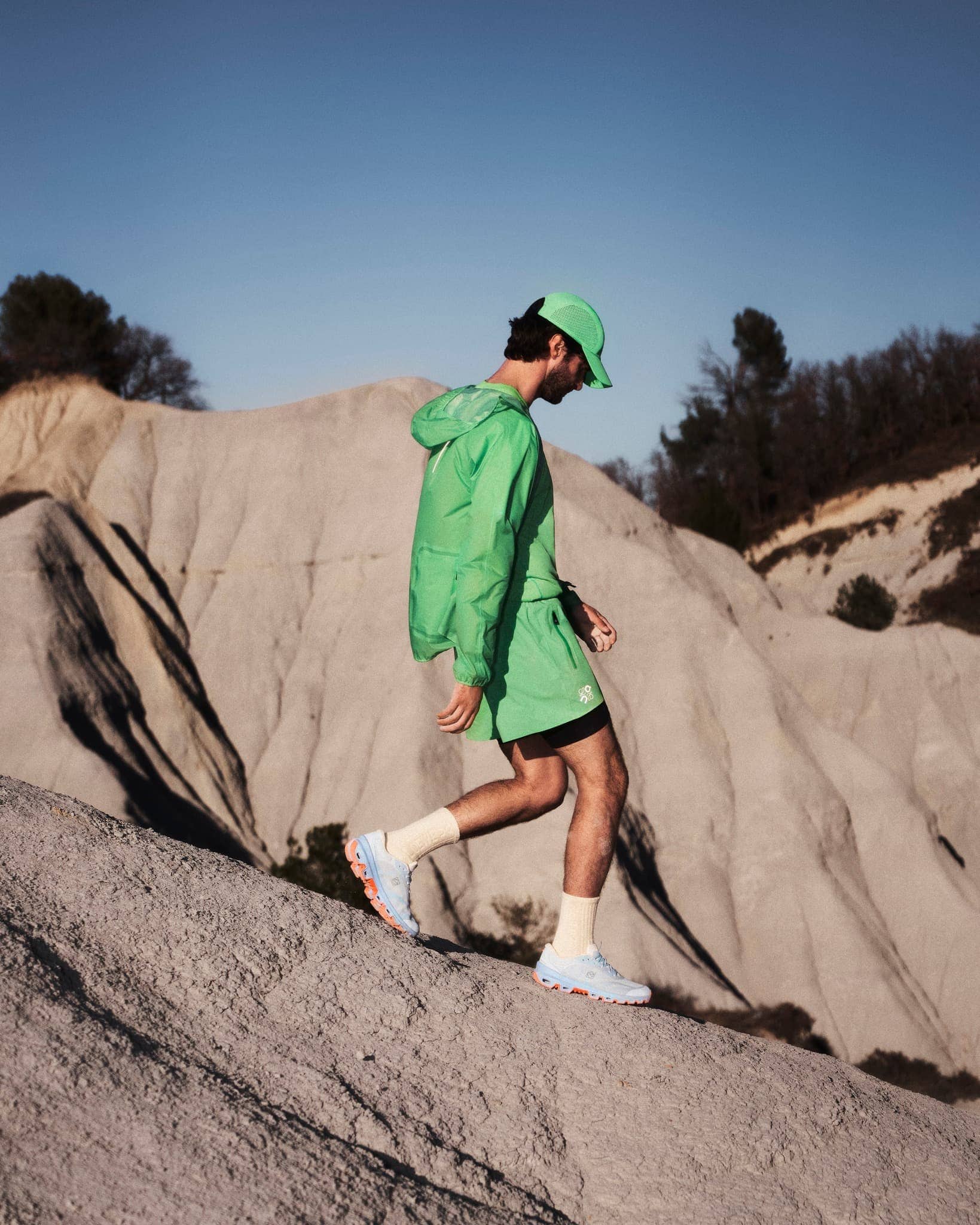 Man wearing a green jacket and cap walking down a rocky dirt slope under a clear sky