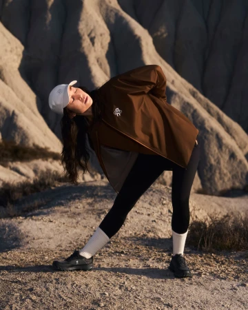 Woman in brown jacket and white cap stretching against a rugged desert landscape