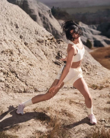 Woman in athletic crop top and shorts posing dynamically on a rugged sand dune landscape outdoors under natural daylight.