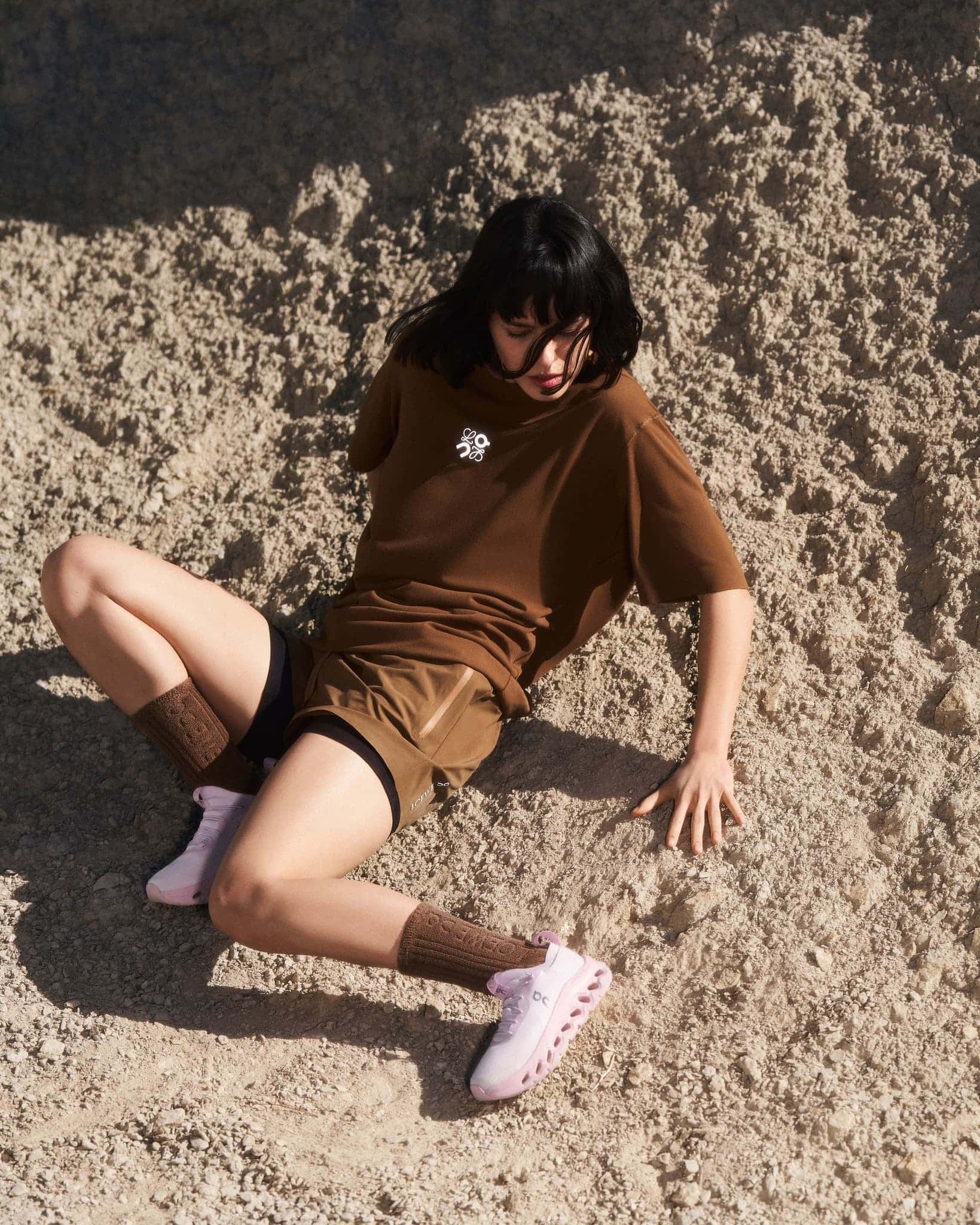 Woman with dark hair wearing brown t-shirt and shorts seated on sandy terrain for Loewe Spring 2025 campaign.