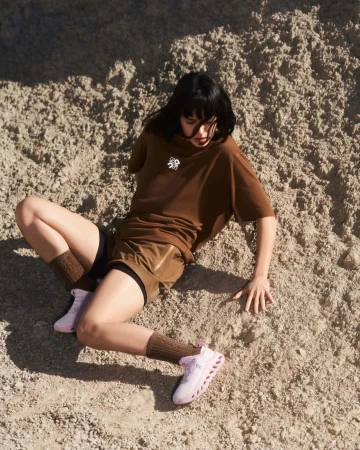 Woman with dark hair wearing brown t-shirt and shorts seated on sandy terrain for Loewe Spring 2025 campaign.