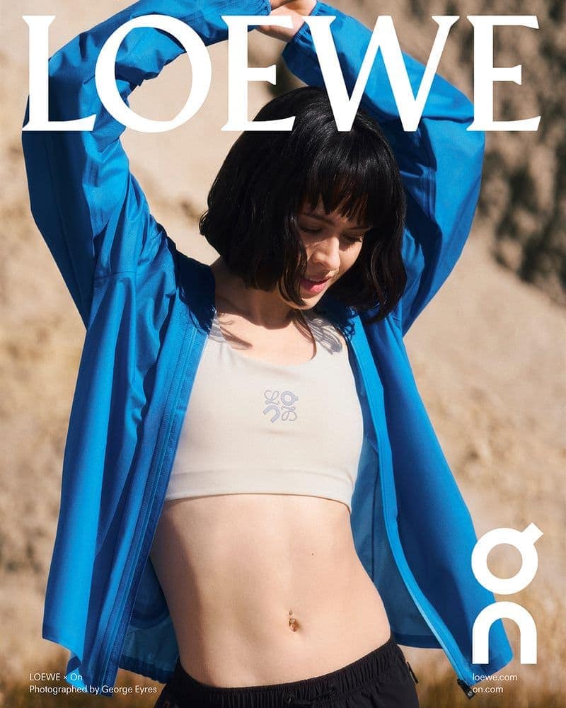 Woman in blue zip jacket and white sports bra stretching arms overhead outdoors