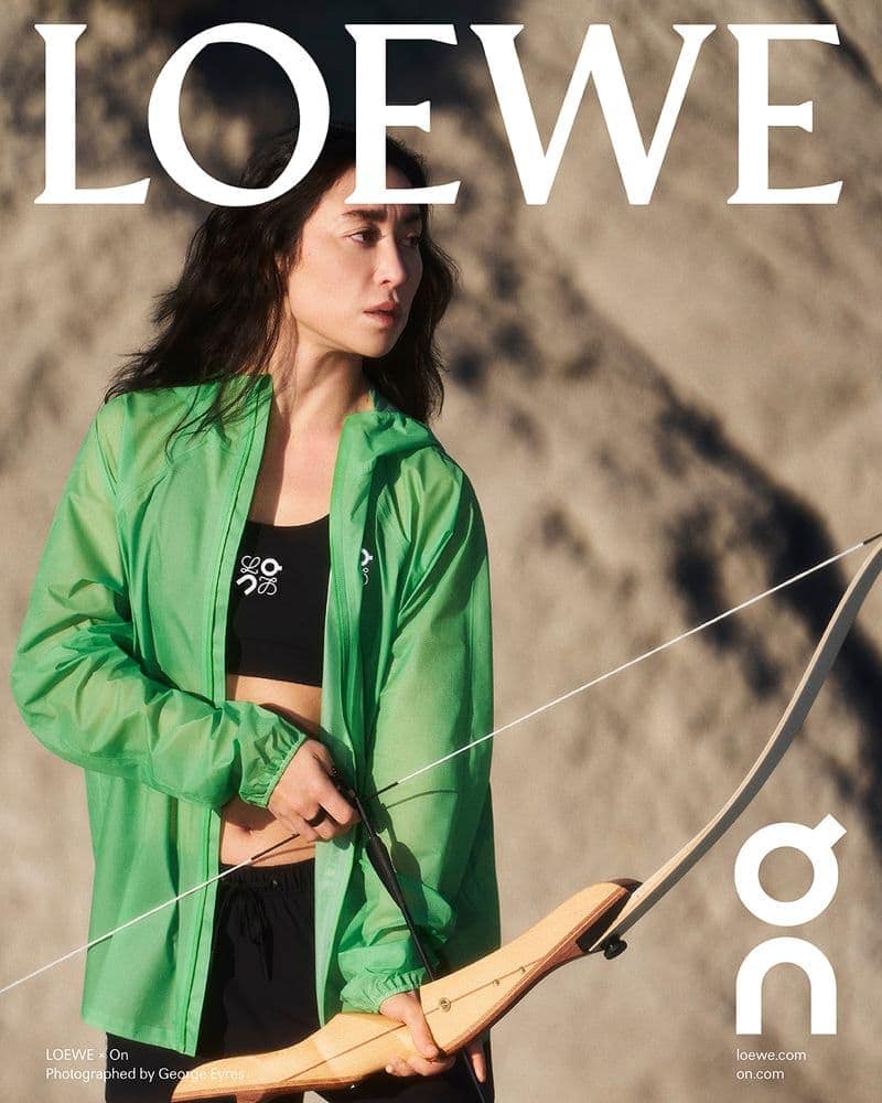 Woman wearing a green zip jacket holding a wooden bow against a sandy dunes background for Loewe.