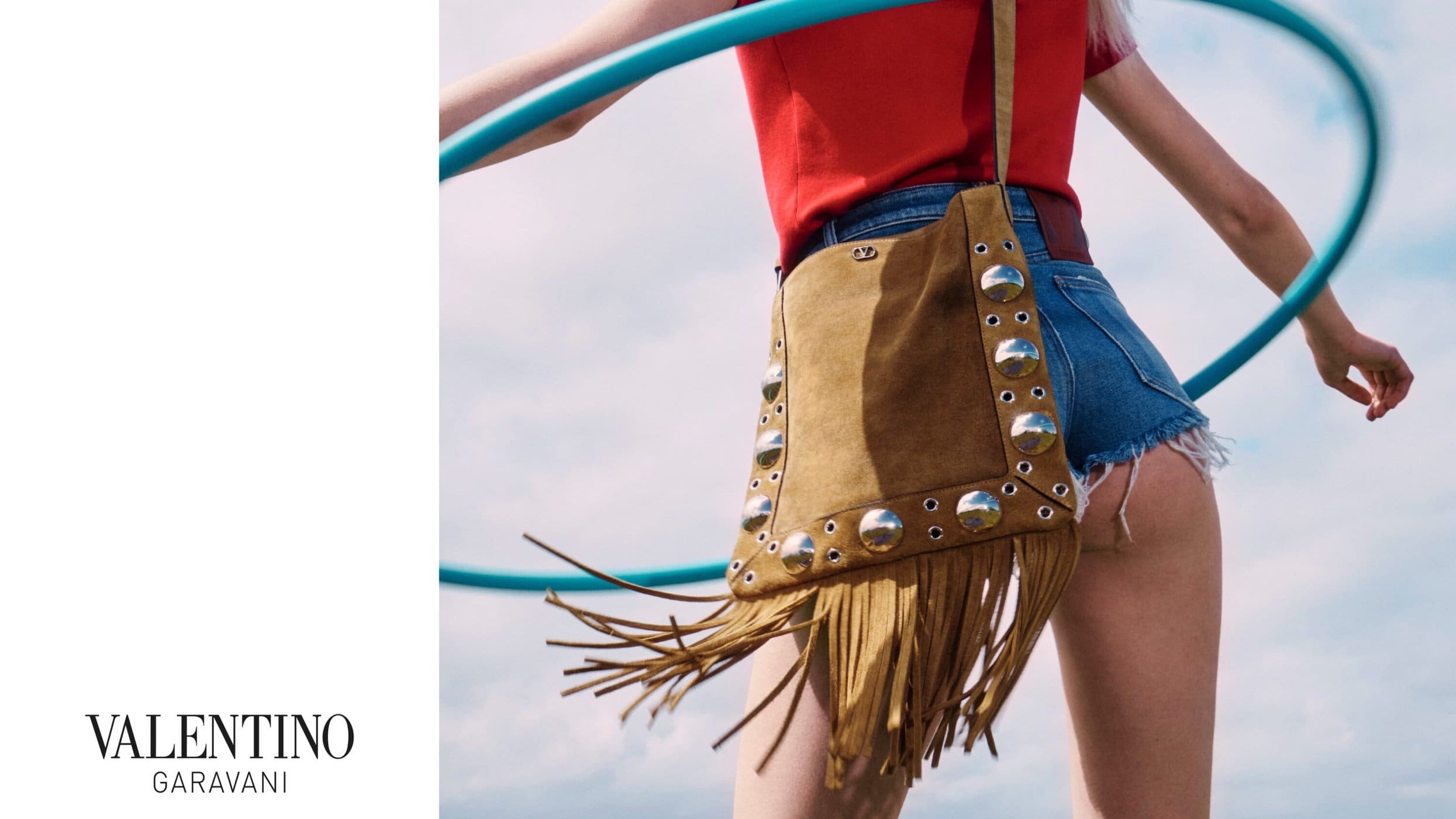 Model wearing denim shorts and a red top holding a hula hoop with a suede fringe bag for Valentino