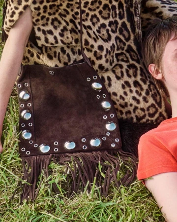 Brown suede fringed shoulder bag with metal grommets and studs resting on green grass next to a model wearing a leopard print garment.
