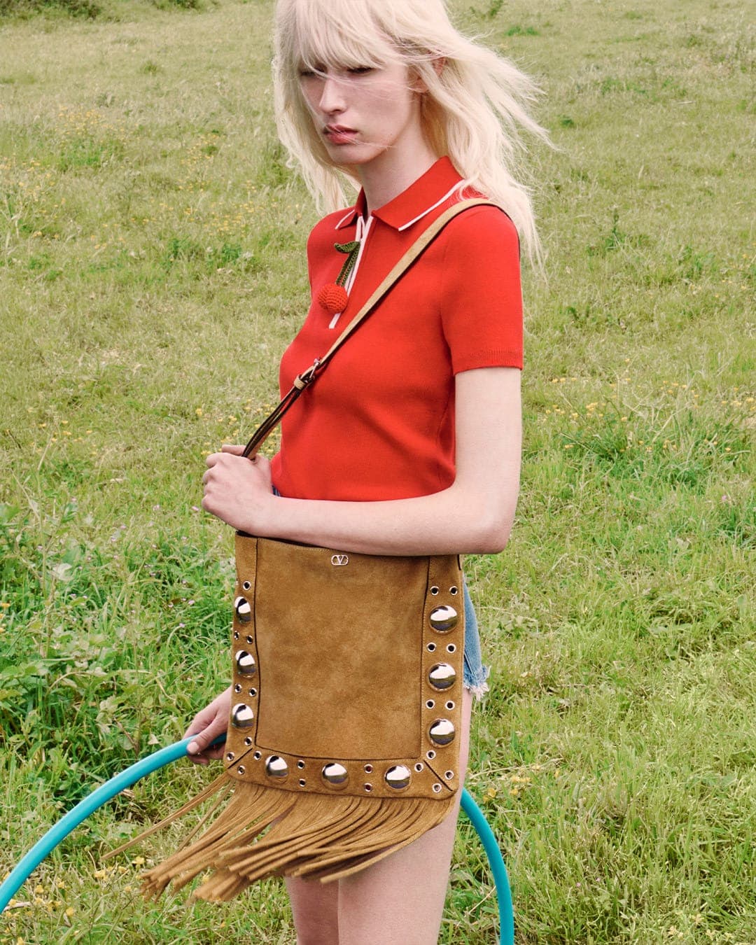 Model with blonde hair wearing a red polo shirt holding a brown fringed suede bag outdoors in a grassy field