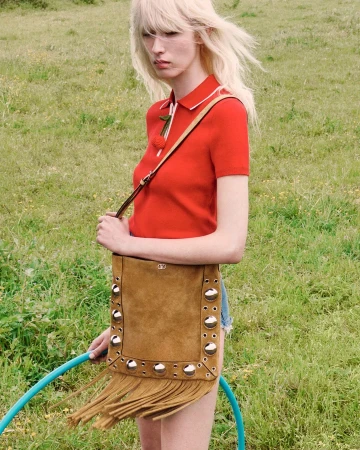 Model with blonde hair wearing a red polo shirt holding a brown fringed suede bag outdoors in a grassy field