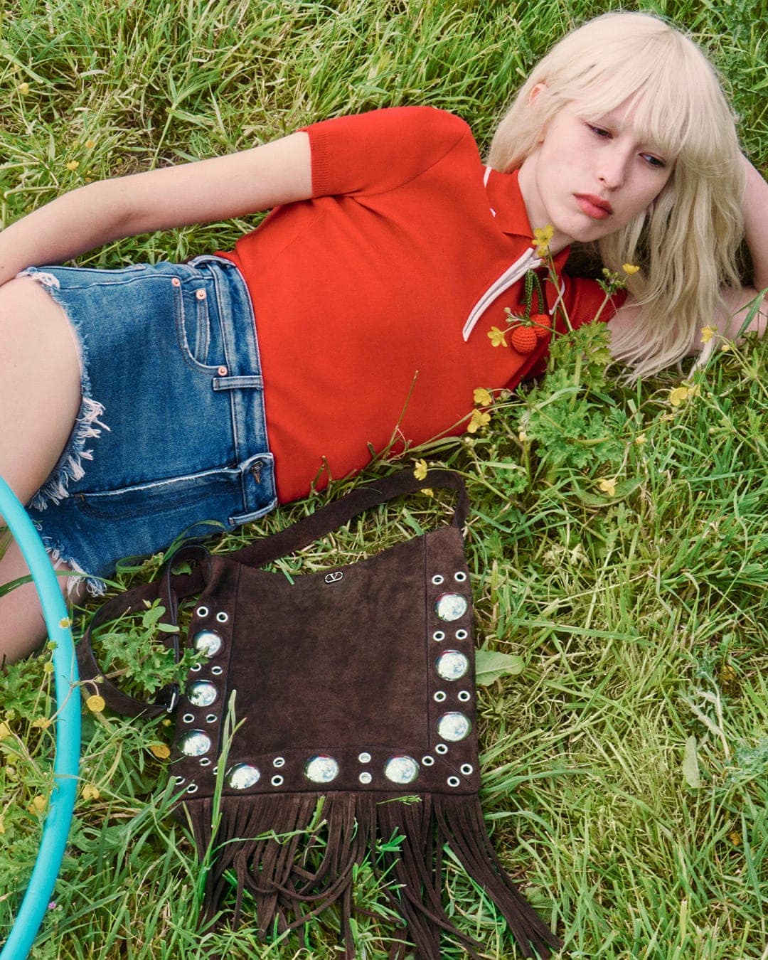 Blonde female model wearing red polo shirt and denim shorts lying in tall green grass with dark brown suede fringe bag