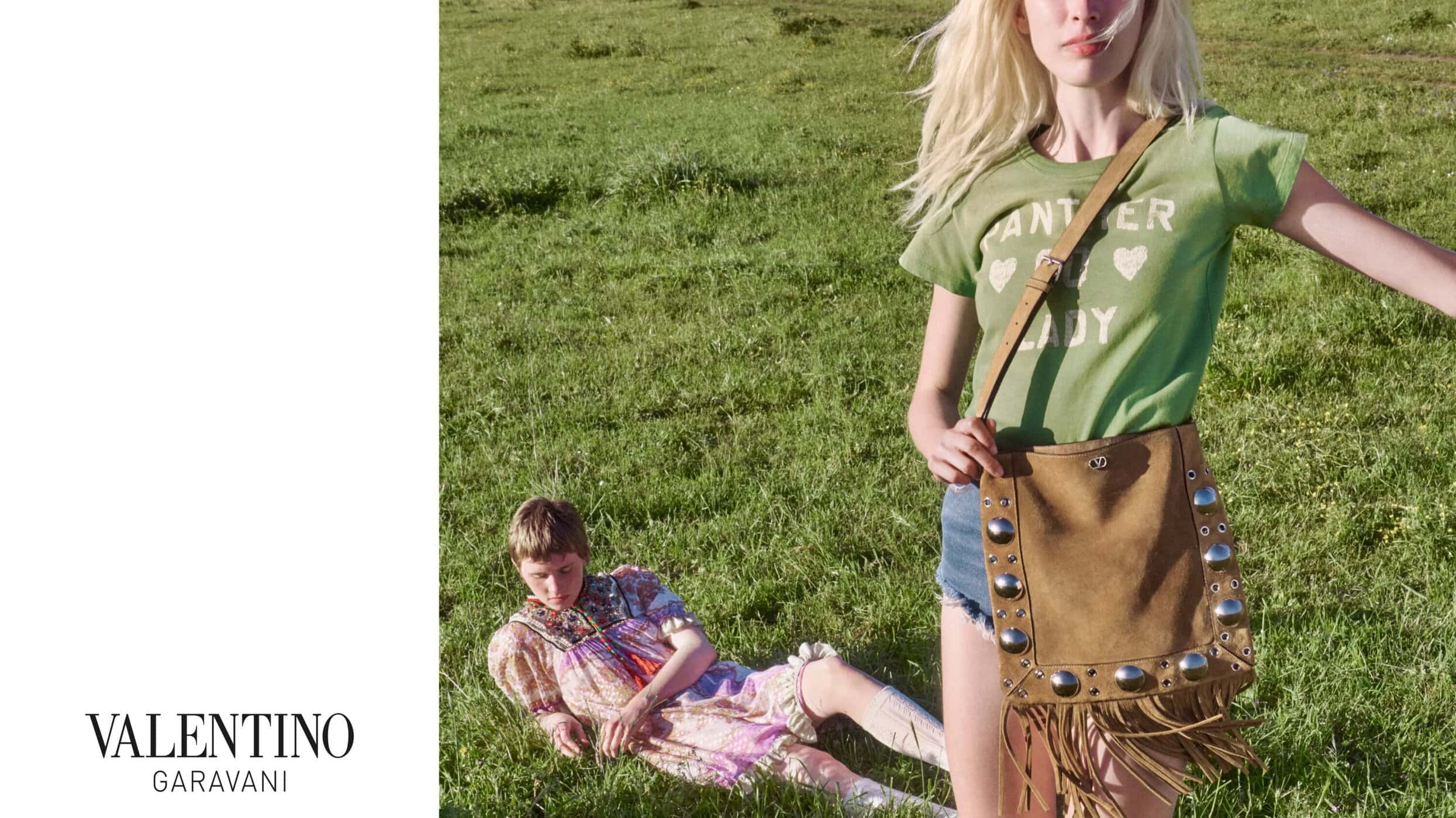 Model standing in a green t-shirt and denim shorts holding a brown fringed shoulder bag in a grassy meadow with another model lying down