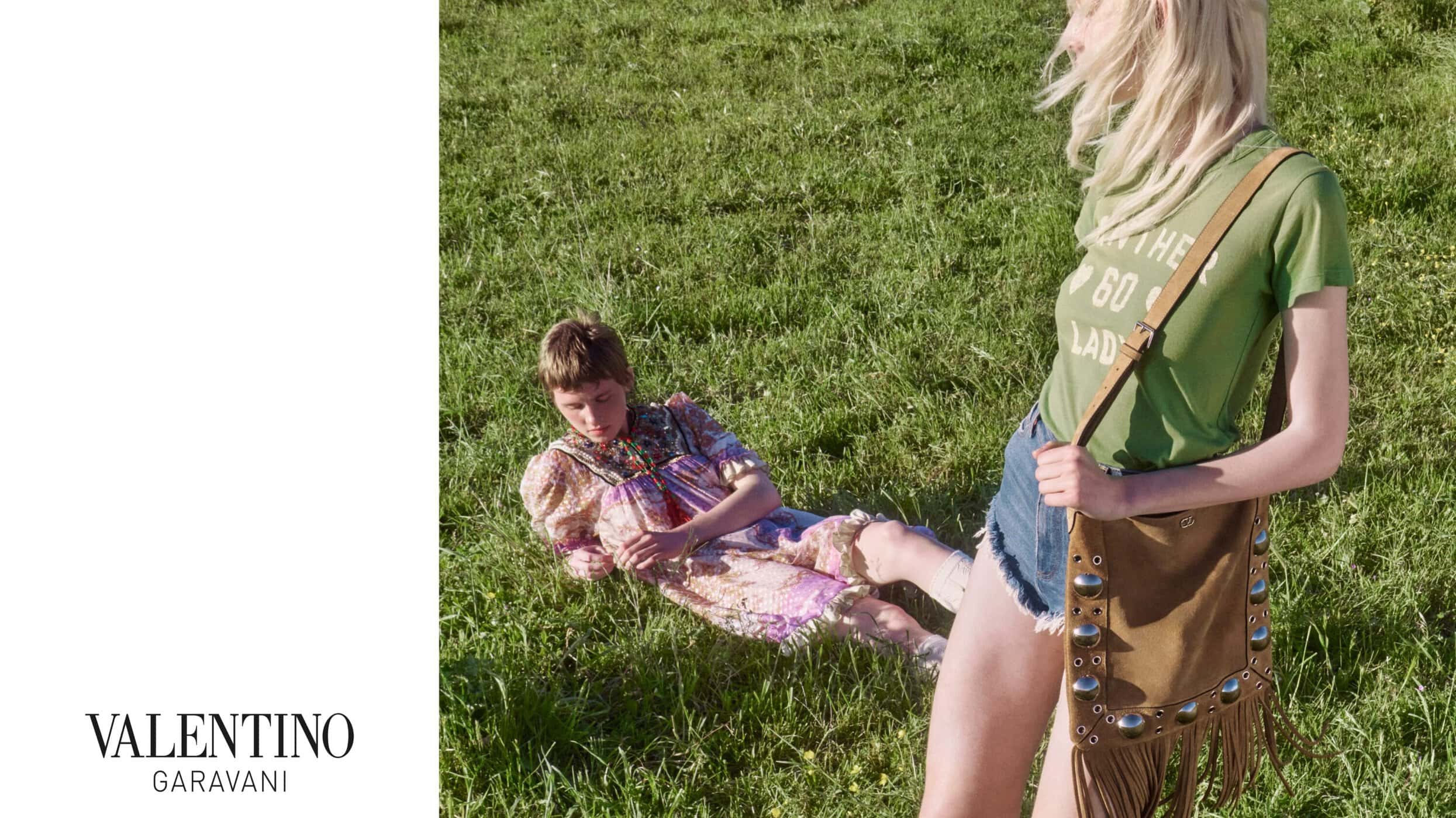 Two models on a grassy meadow, one standing wearing a green t-shirt and holding a brown fringed shoulder bag with metal studs.