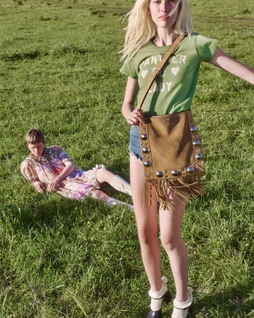 Model standing in a green meadow wearing a green t-shirt and holding a brown fringed studded shoulder bag with another person seated behind.