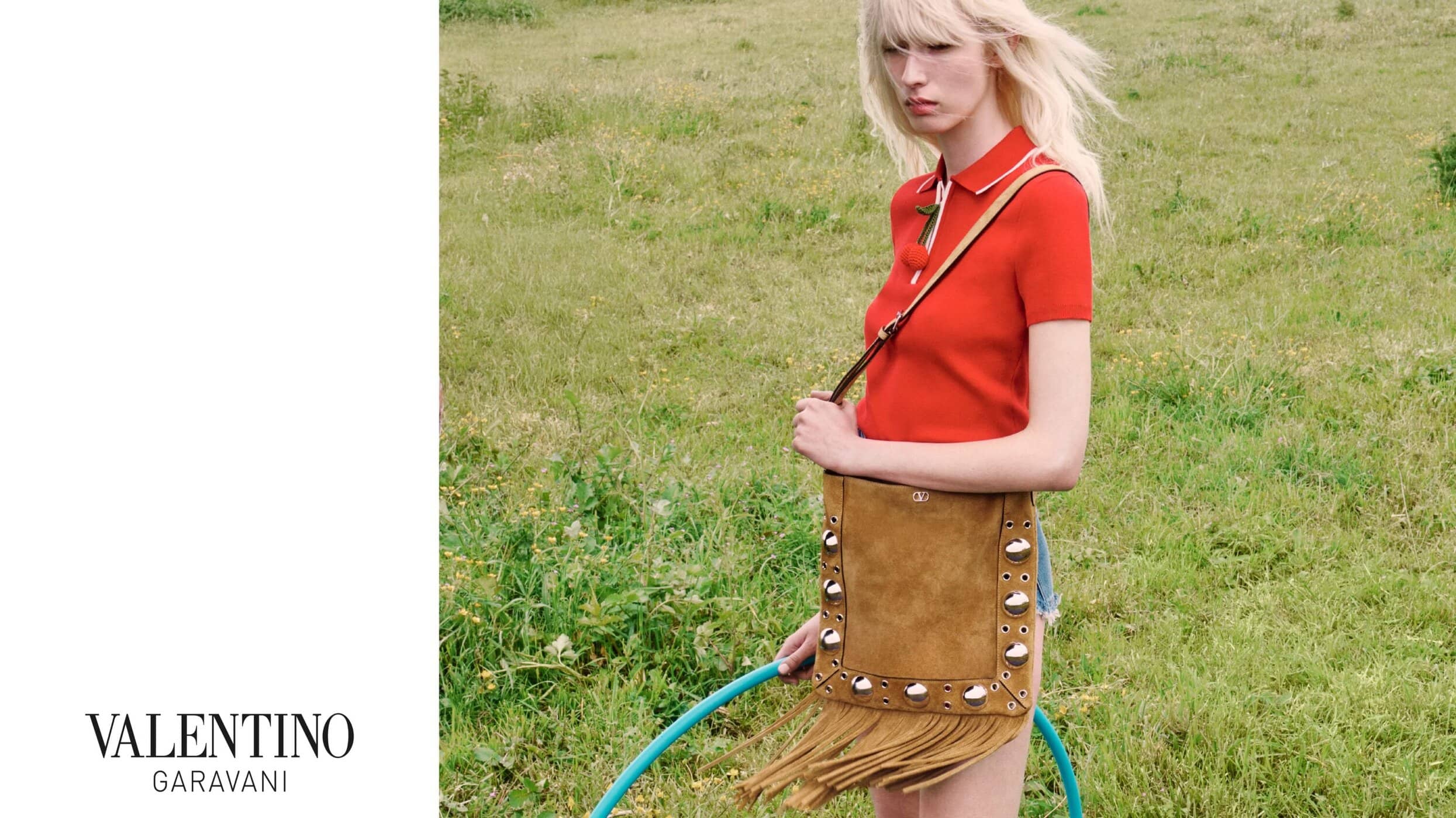 Model in red polo shirt holding fringed tan suede shoulder bag against grassy meadow with white branding panel on left