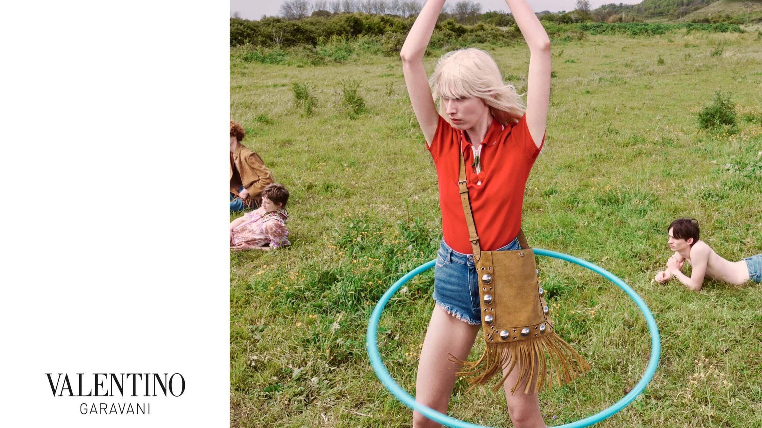 Blonde woman wearing a red polo shirt and denim shorts holding a fringed suede bag while spinning a blue hula hoop in a grassy meadow