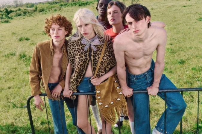Group of five models leaning on a black metal barrier in a grassy meadow, featuring a Valentino fringed suede handbag