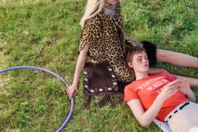 Models lying in a grassy field, one wearing a leopard print cape and holding a fringed brown suede handbag by Valentino.