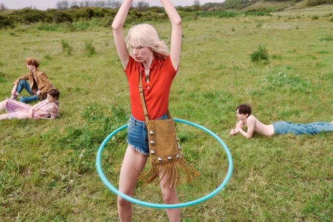 Model in red polo shirt and denim shorts holding a fringed suede bag while spinning a hula hoop in a grassy field with other models.