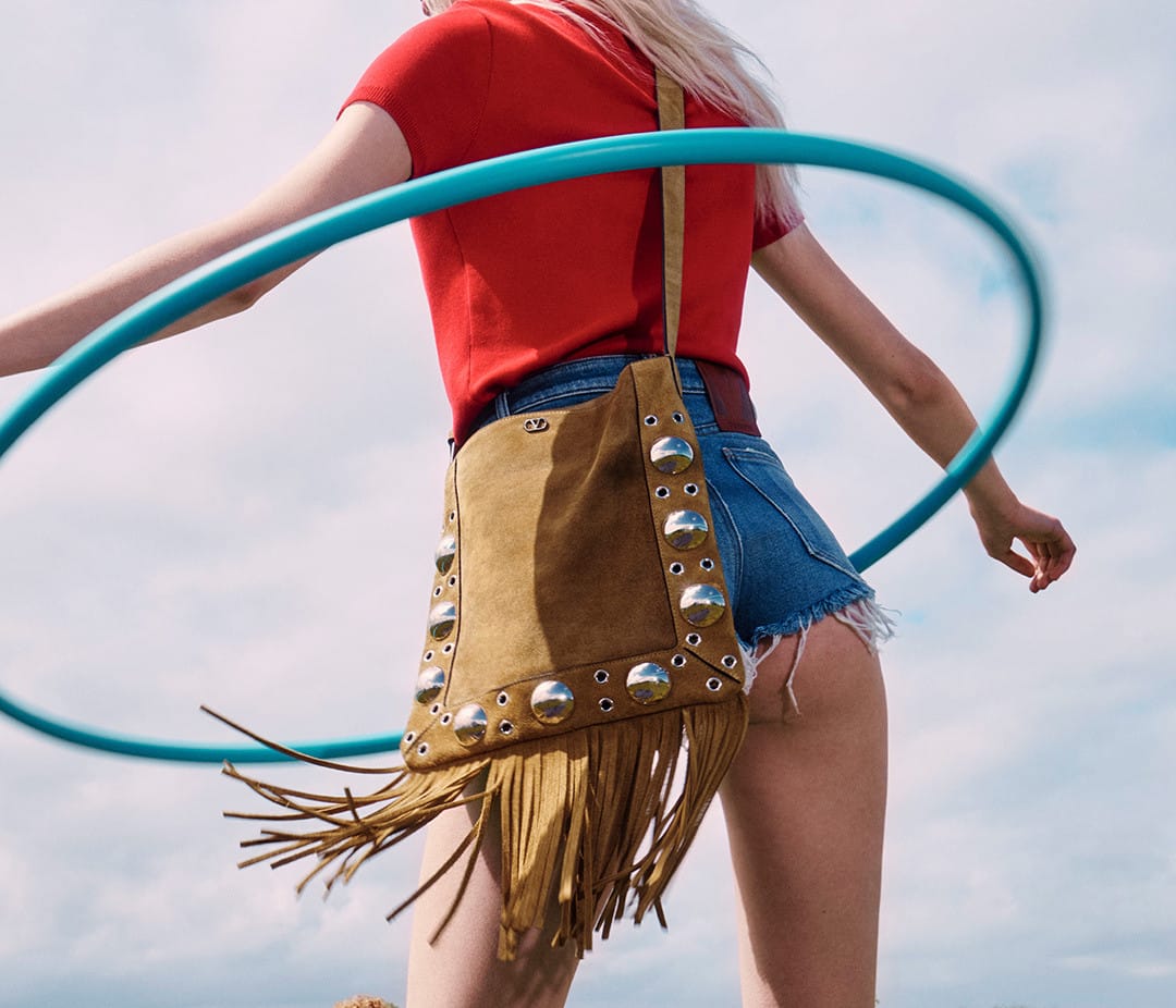 Model wearing a red t-shirt and denim shorts with a fringed suede bag while spinning a blue hula hoop against an overcast sky.