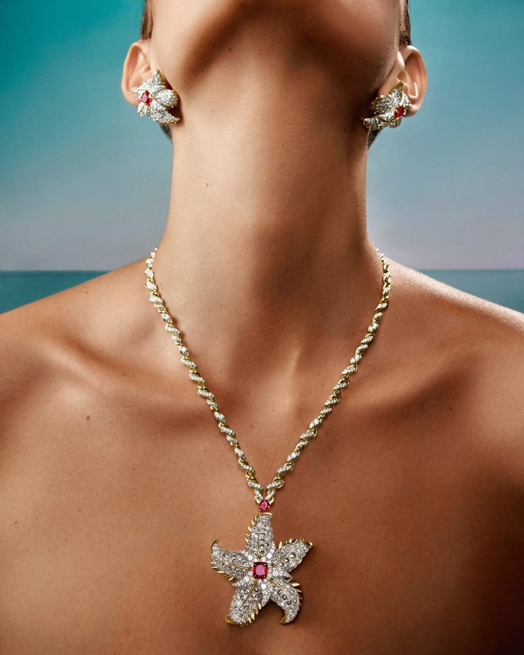 Close-up portrait of a model wearing diamond and ruby starfish necklace and matching earrings against a blue background.