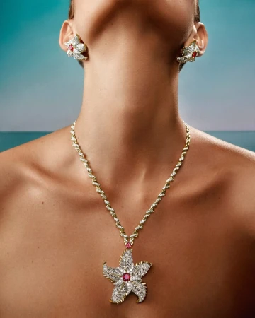 Close-up portrait of a model wearing diamond and ruby starfish necklace and matching earrings against a blue background.