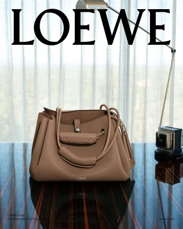 Loewe leather handbag resting on a dark polished wooden table with a metal desk lamp and sheer curtain background.