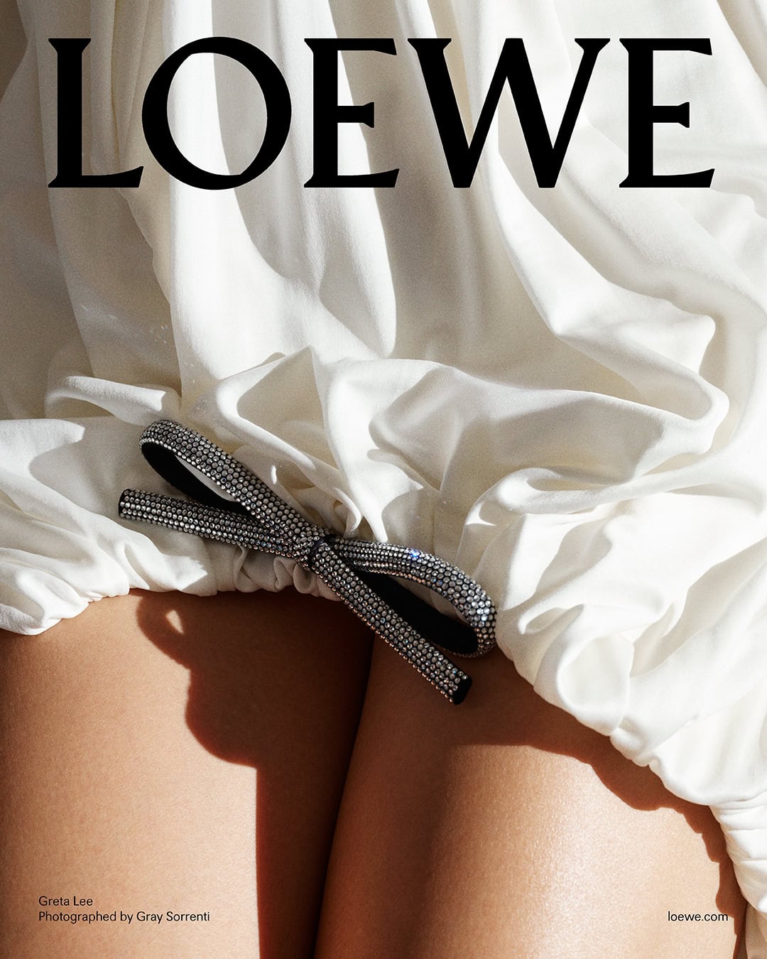 Close-up of draped white fabric with an embellished crystal bow resting on bare legs for a Loewe Pre-Fall 2025 campaign image.