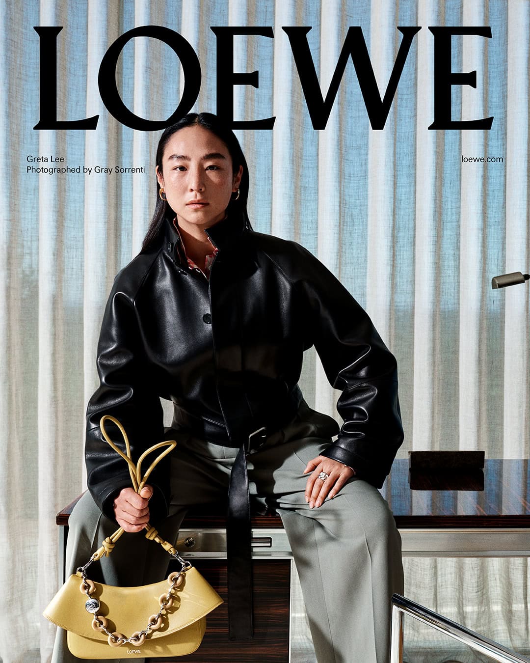 Greta Lee wearing a black leather jacket and grey trousers holding a yellow handbag against textured curtains for Loewe Pre-Fall 2025.