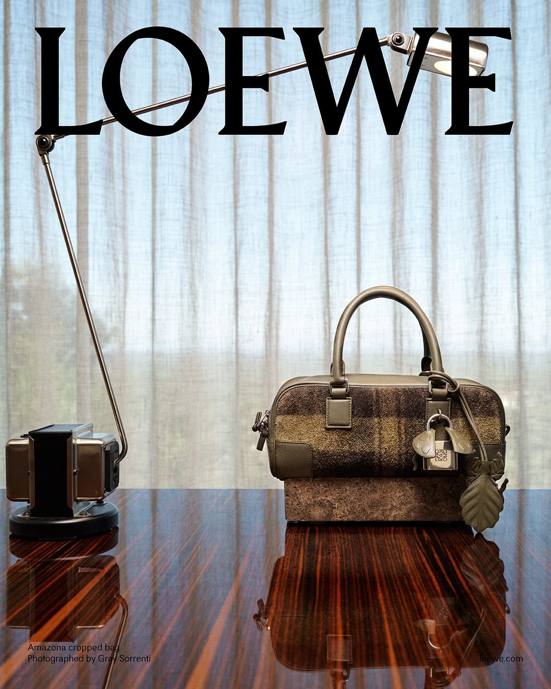 Loewe campaign image featuring a structured handbag and a sleek metal desk lamp on a polished wooden table against a sheer curtain background.
