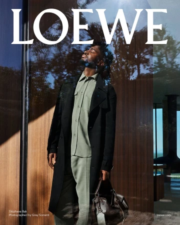 Stéphane Bak wearing a sage green shirt and trousers under a black coat holding a leather bag against a wood paneled interior wall