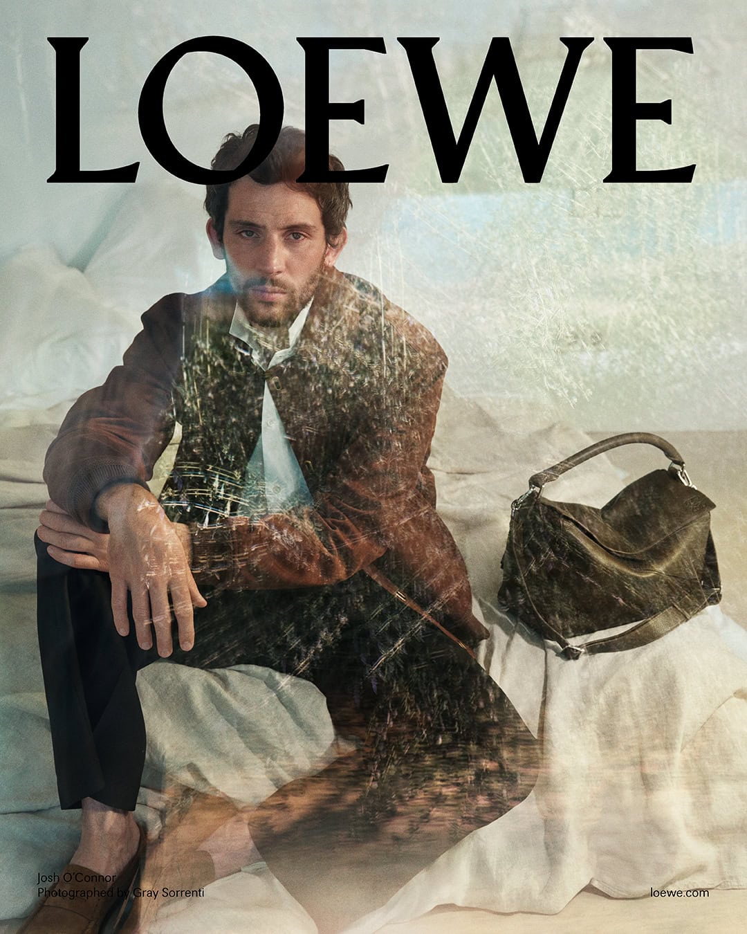 Josh O'Connor seated on a rumpled white textile surface beside a leather handbag, with a double exposure effect.