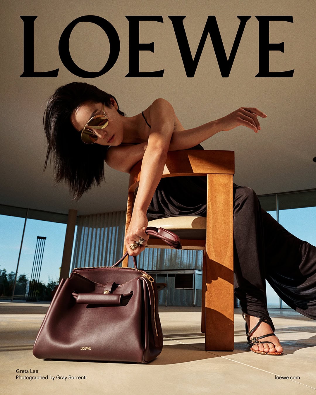 Greta Lee wearing sunglasses and black clothing, leaning over a wooden chair while holding a burgundy leather handbag in a bright interior.